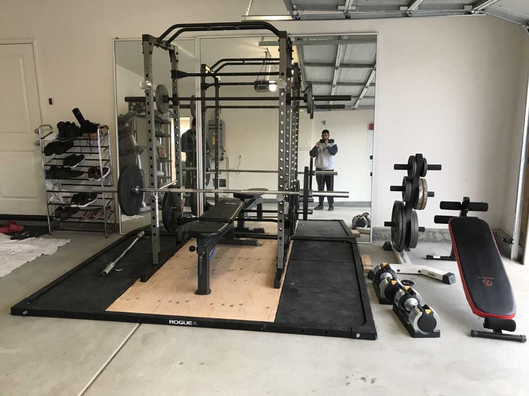 A home gym in a garage with a squat rack, bench, barbells with weights, adjustable dumbbells, an adjustable bench, weight plates on a rack, and a large wall mirror reflecting a person taking a photo.
