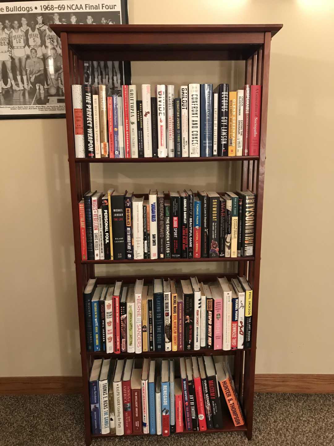 A wooden bookshelf with four shelves filled with neatly arranged books. The books are of various sizes and colors. A framed photo and part of a basketball poster hang on the beige wall behind the shelf.