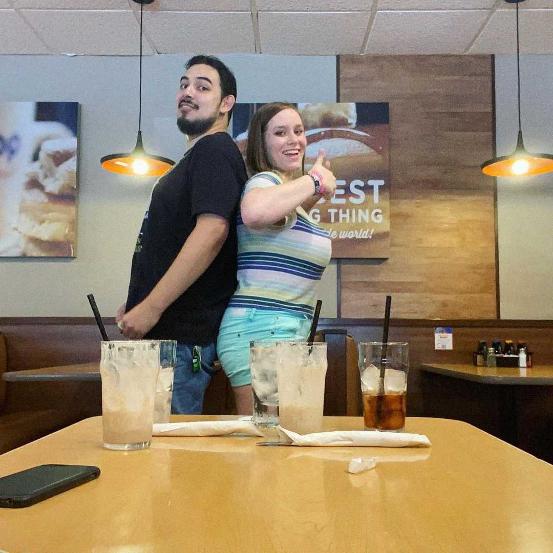 A man and a woman stand back-to-back, smiling and posing with thumbs up, in a restaurant booth. Four drinks and napkins are on the table in front of them. Warm lighting and framed photos decorate the background.