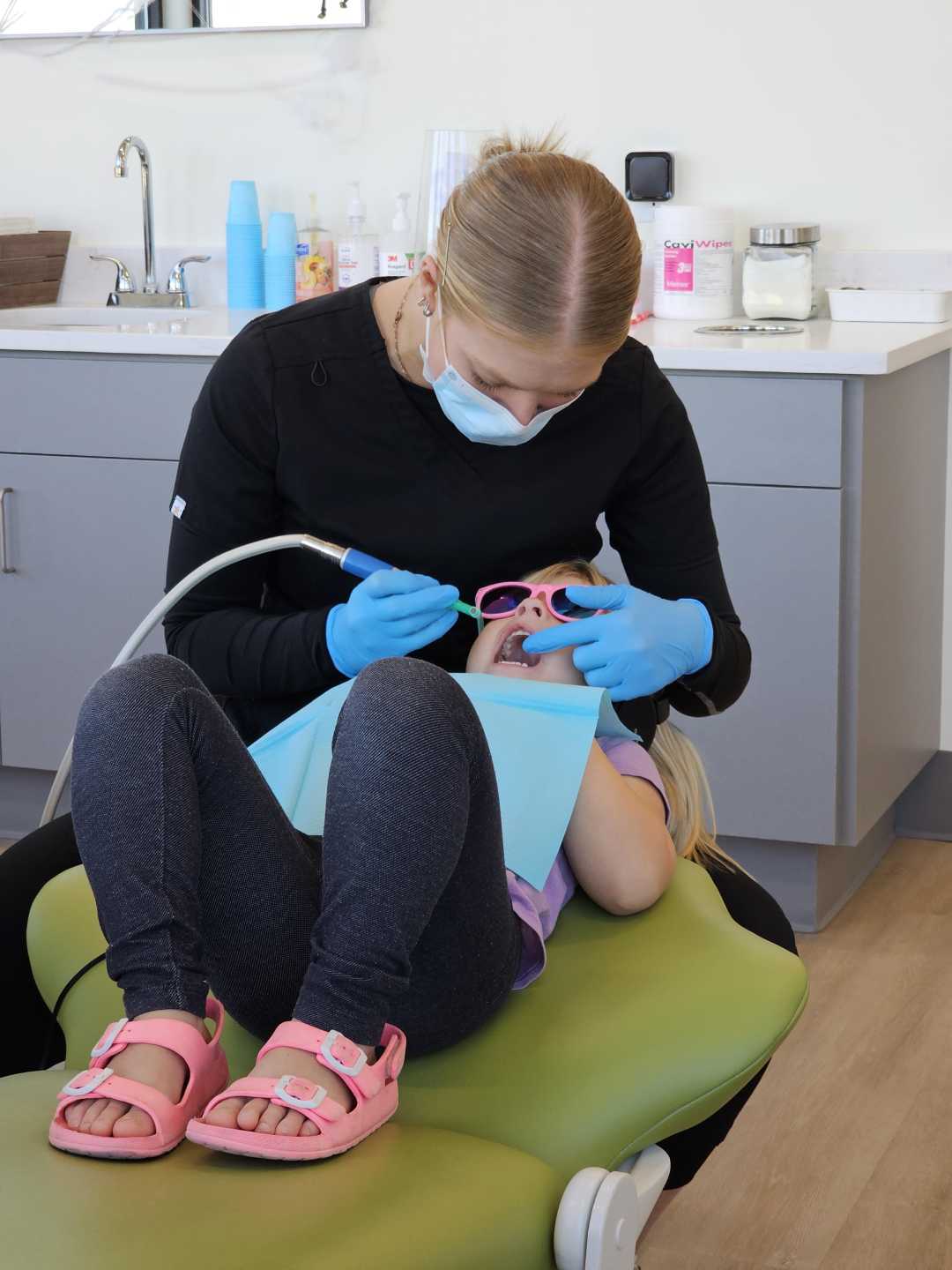 A dental hygienist in gloves and a mask cleans a young girl's teeth. The girl, wearing pink sandals and sunglasses, lies back in a green dental chair in a modern clinic.