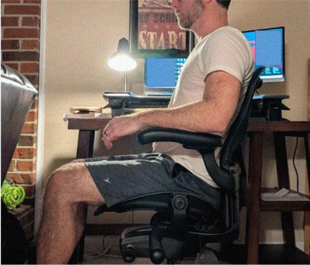 A man in a t-shirt and shorts sits upright in an office chair at a desk with a lamp, two laptops, and a monitor, in a home office setting.