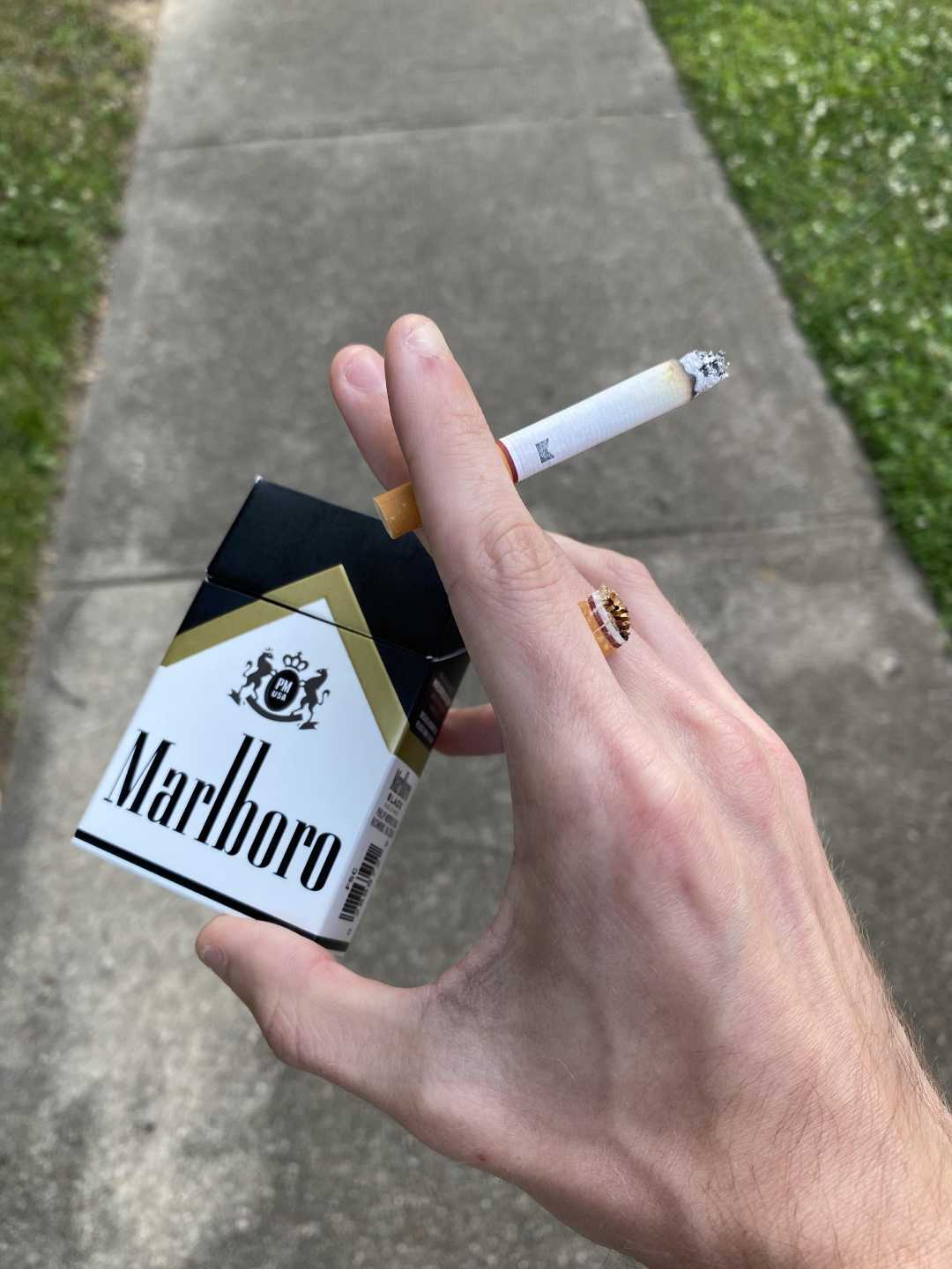 A person holding a Marlboro cigarette pack and a lit cigarette between their fingers, standing on a sidewalk with grass visible to the side.