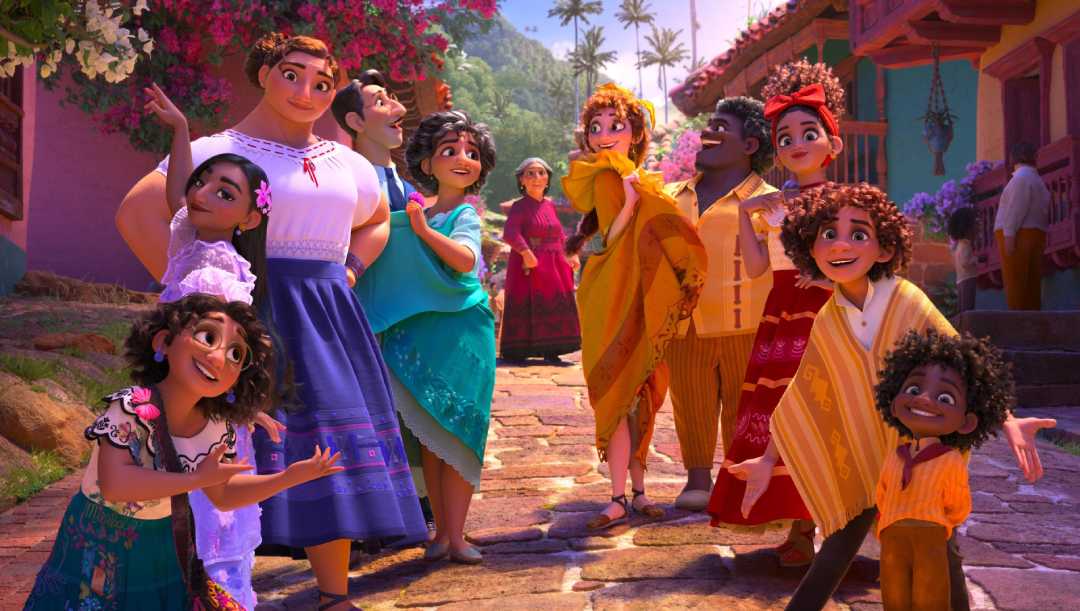 A group of animated characters from the movie "Encanto" stand together, smiling and posing on a colorful cobblestone street, surrounded by vibrant houses and flowers in the background.