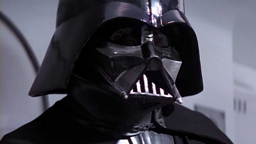 A person in a black, shiny helmet and mask with a triangular mouthpiece and dark cape, standing indoors against a white background.