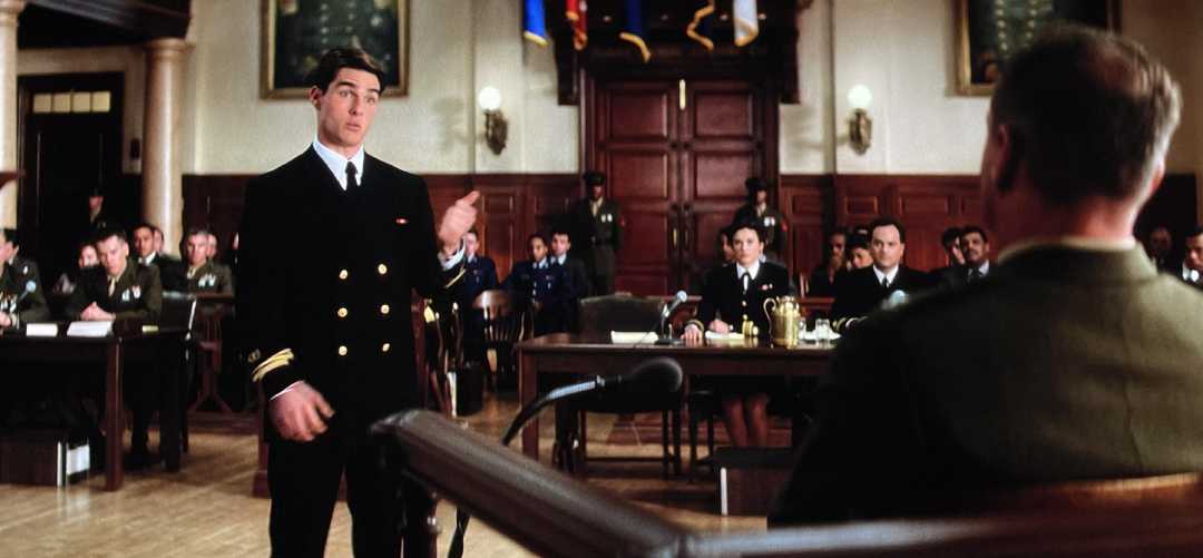 A man in a dark naval uniform stands and speaks in a courtroom, addressing a seated man in military attire as others look on attentively from benches in the background.