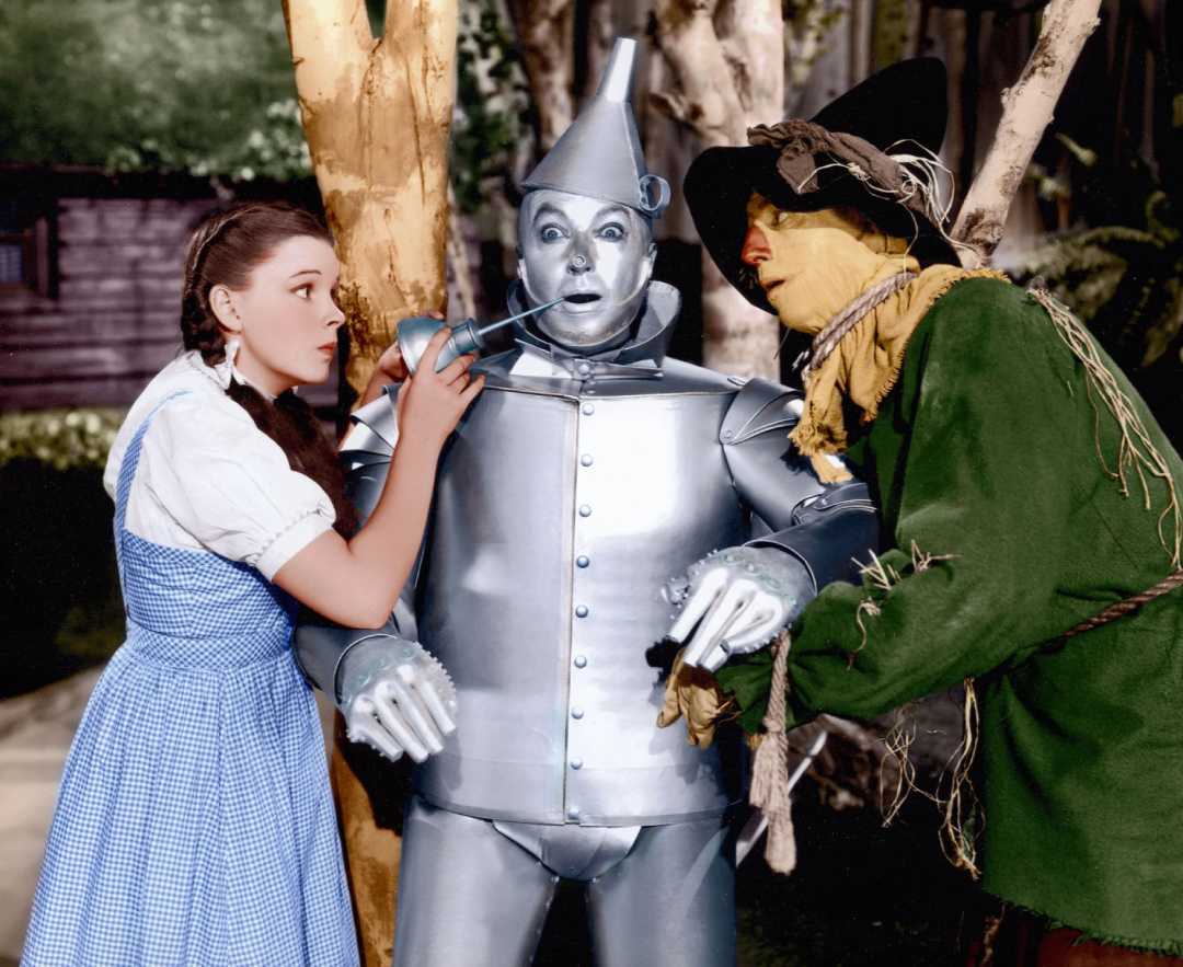 A young woman in a blue dress oils the mouth of a silver Tin Man, while a man dressed as a Scarecrow in a green outfit and straw hat stands beside them, both watching closely.