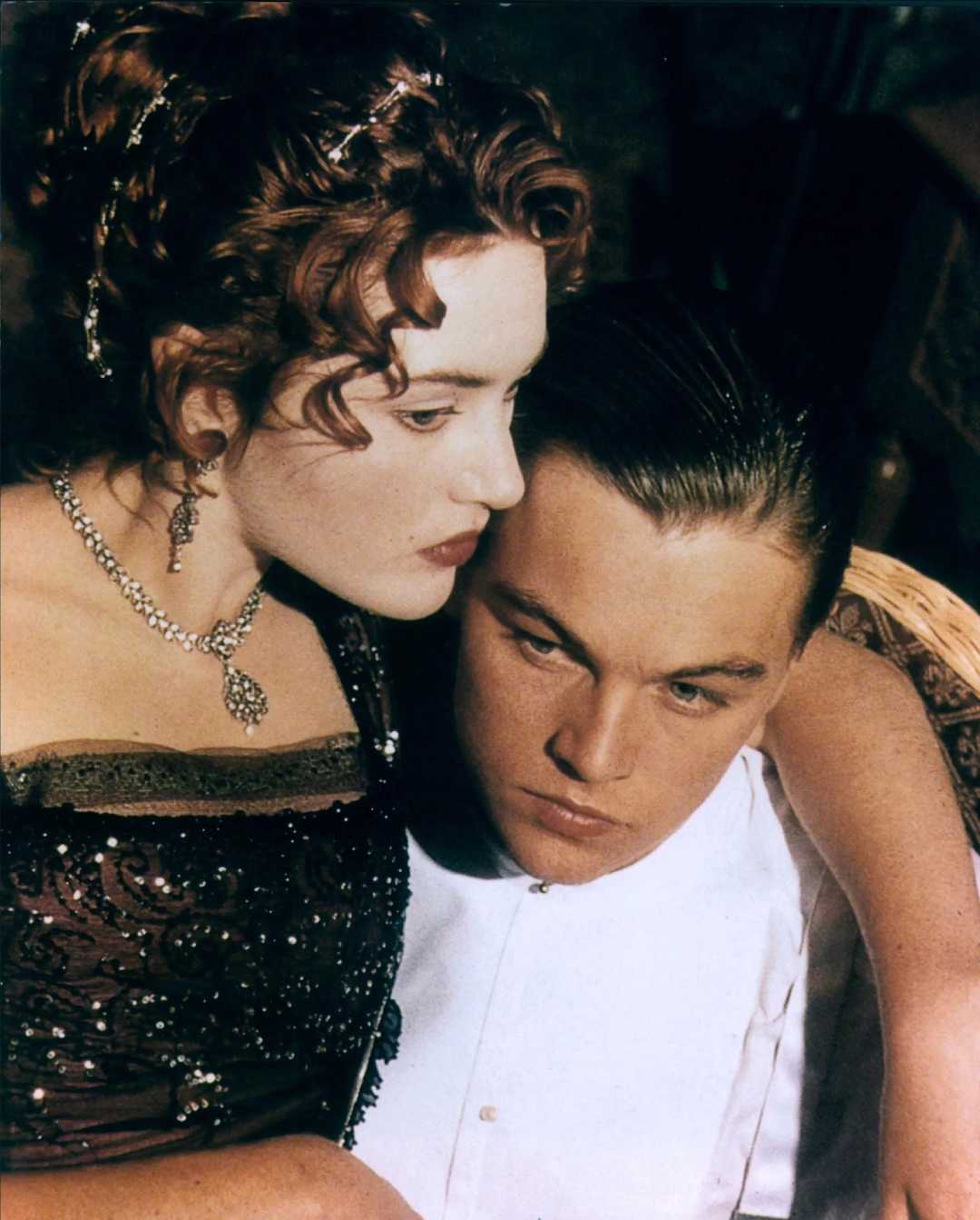A woman in an ornate dress and jewelry leans closely against a man in formal attire. Both appear serious and are looking in the same direction, suggesting a moment of deep connection or contemplation.