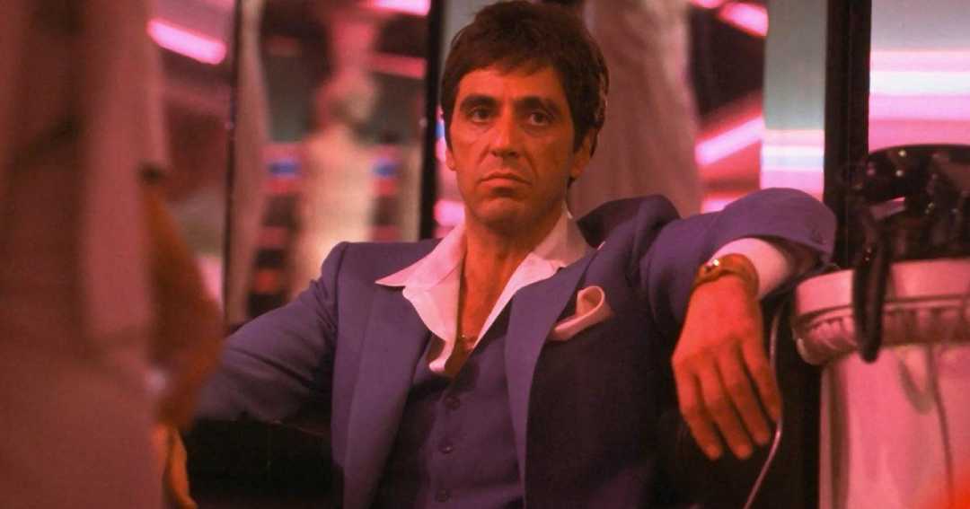 A serious-looking man in a blue suit with an open-collared white shirt sits in a dimly lit room with pink and purple neon lights, leaning back with one arm resting on a chair.
