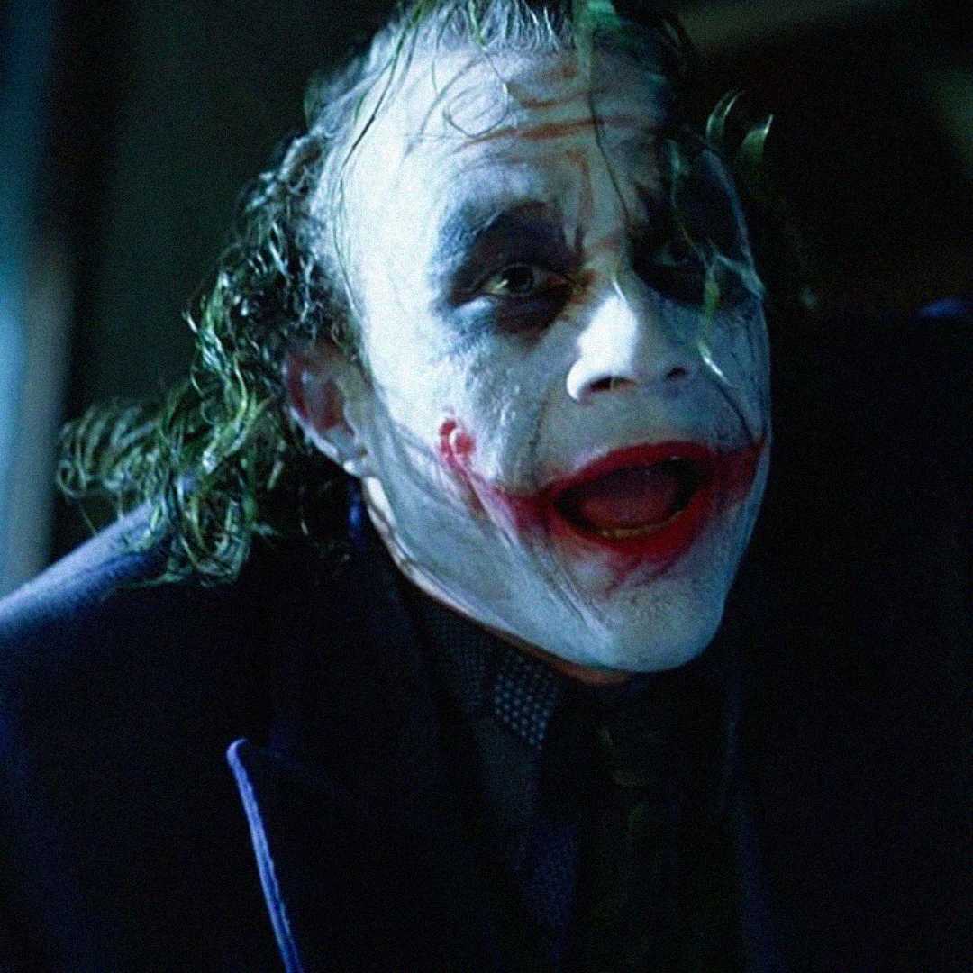 A person with messy green hair and white face paint, red scar-like makeup on the lips, and dark eye makeup, wearing a dark coat, leans forward and looks intensely at the camera.