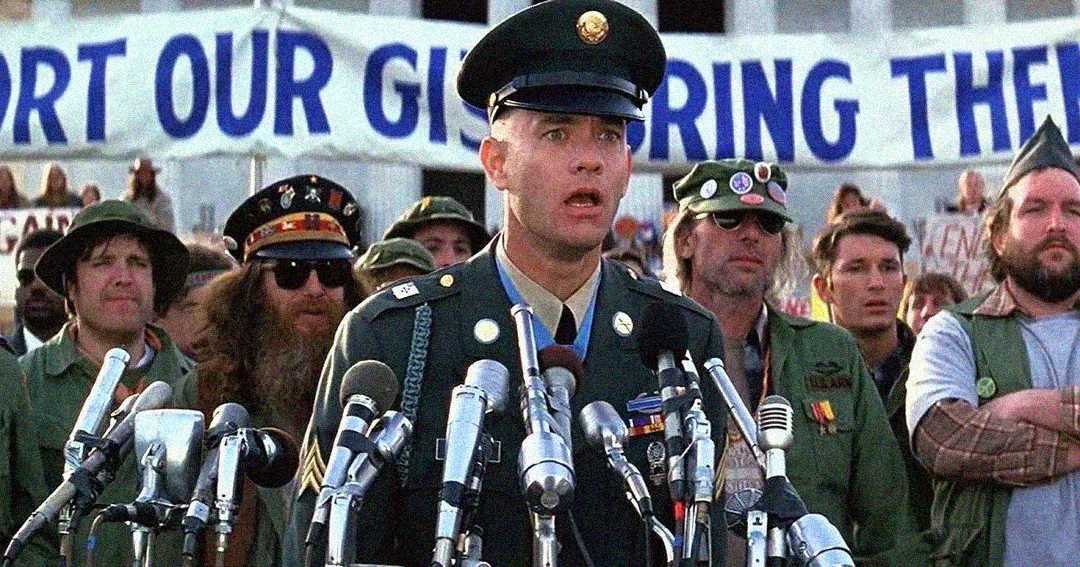 A man in a military uniform stands at a podium with microphones, speaking to a crowd. Behind him, people in casual and military-style clothing stand in front of a large protest banner.