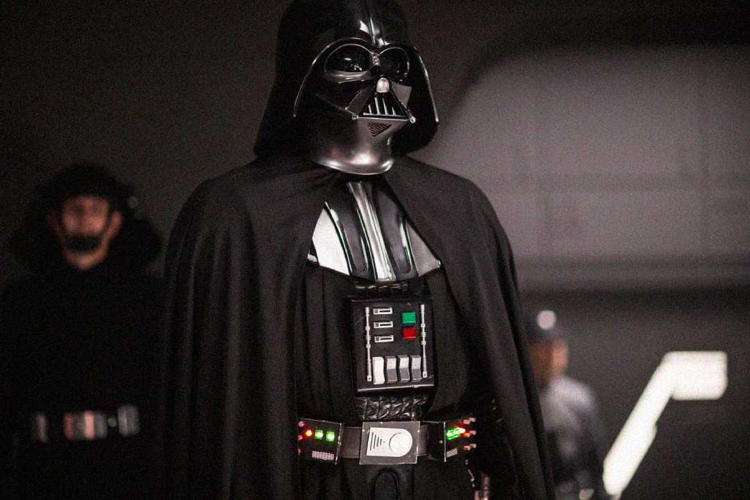A person dressed as Darth Vader from Star Wars stands in a dark room, wearing a black helmet, armor, and cape. Two blurred figures in dark uniforms are visible in the background.