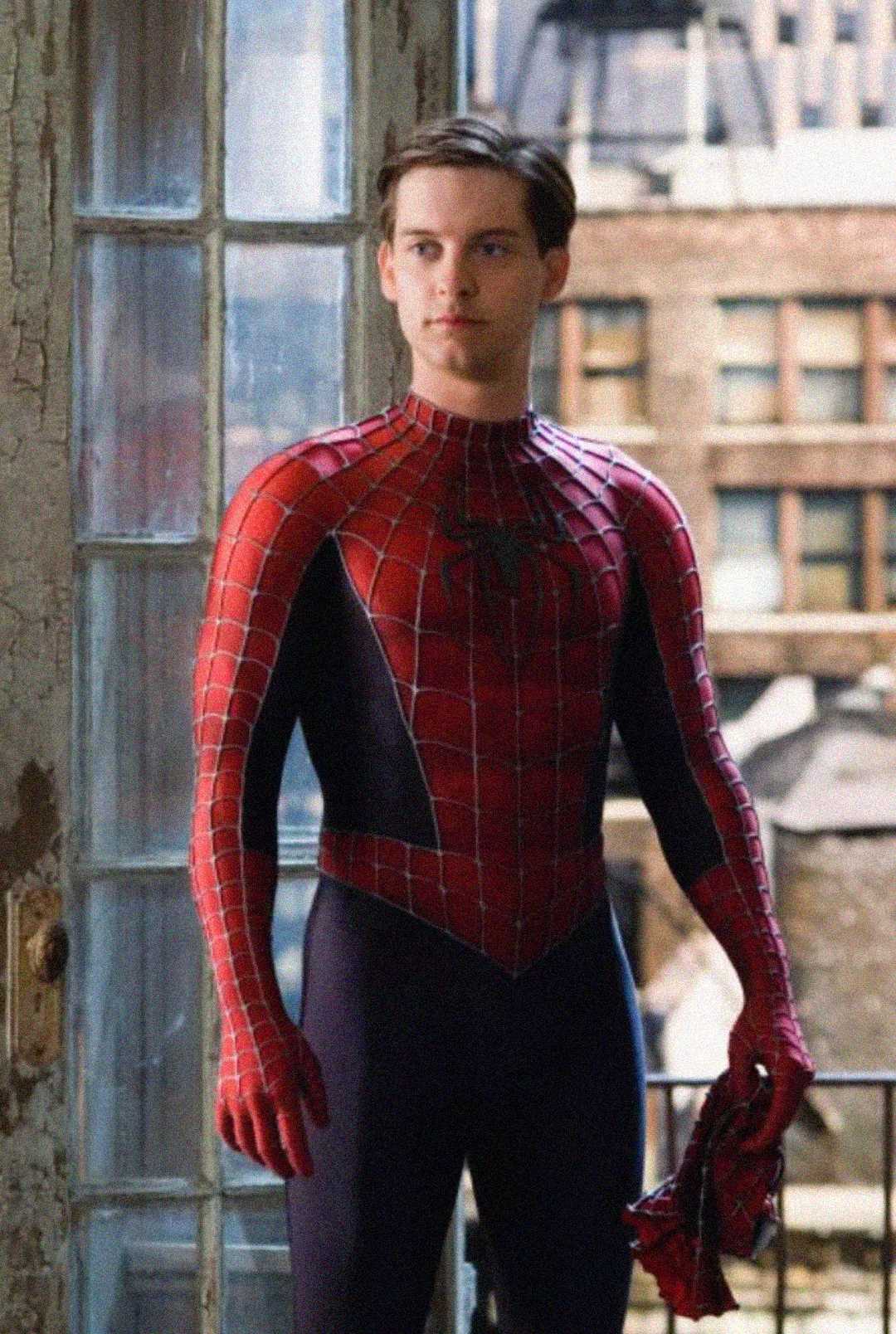 A person wearing a classic red and blue Spider-Man suit stands inside by a glass door, holding the mask in one hand, with city buildings visible through the window behind them.