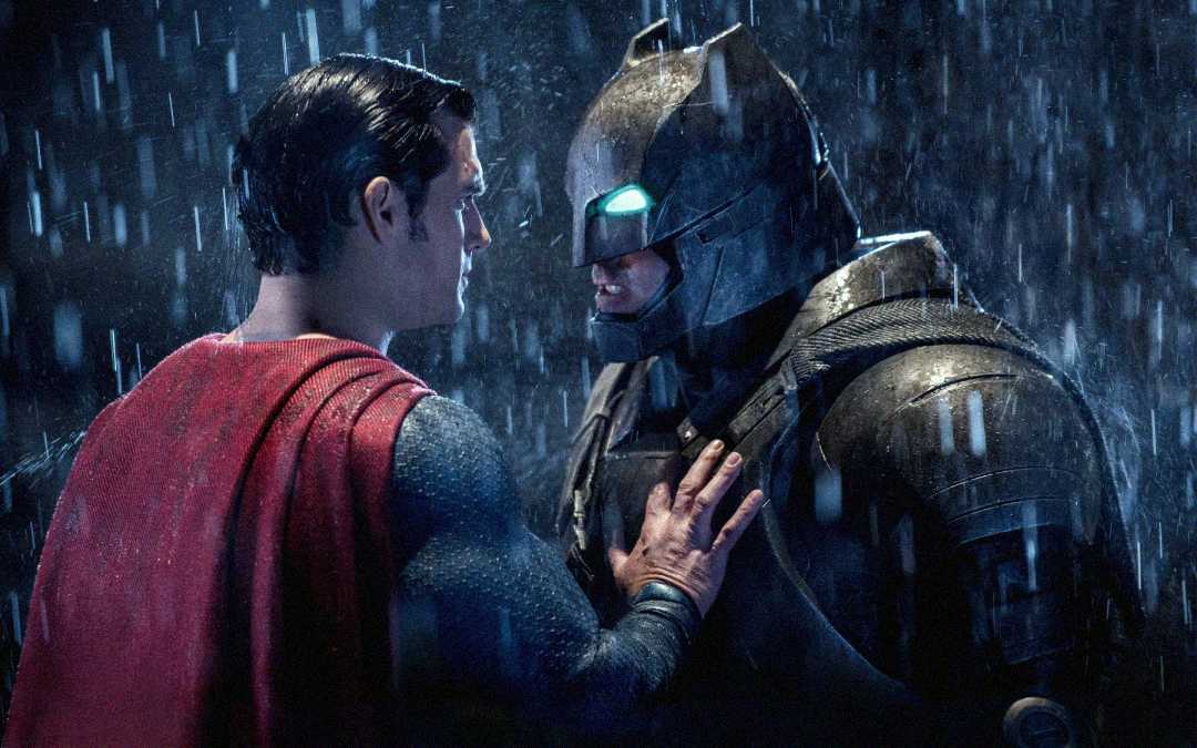 Superman and Batman face each other closely in the rain at night, with Superman’s hand on Batman’s armored chest. Both look intense, and Batman’s eyes glow blue through his mask.