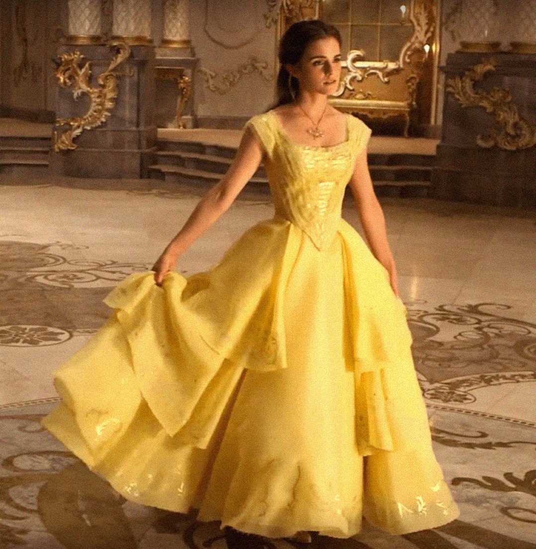 A young woman in a flowing yellow ballgown stands gracefully in an ornate, golden ballroom, holding the sides of her dress and looking to the side. The setting is elegant and grand.