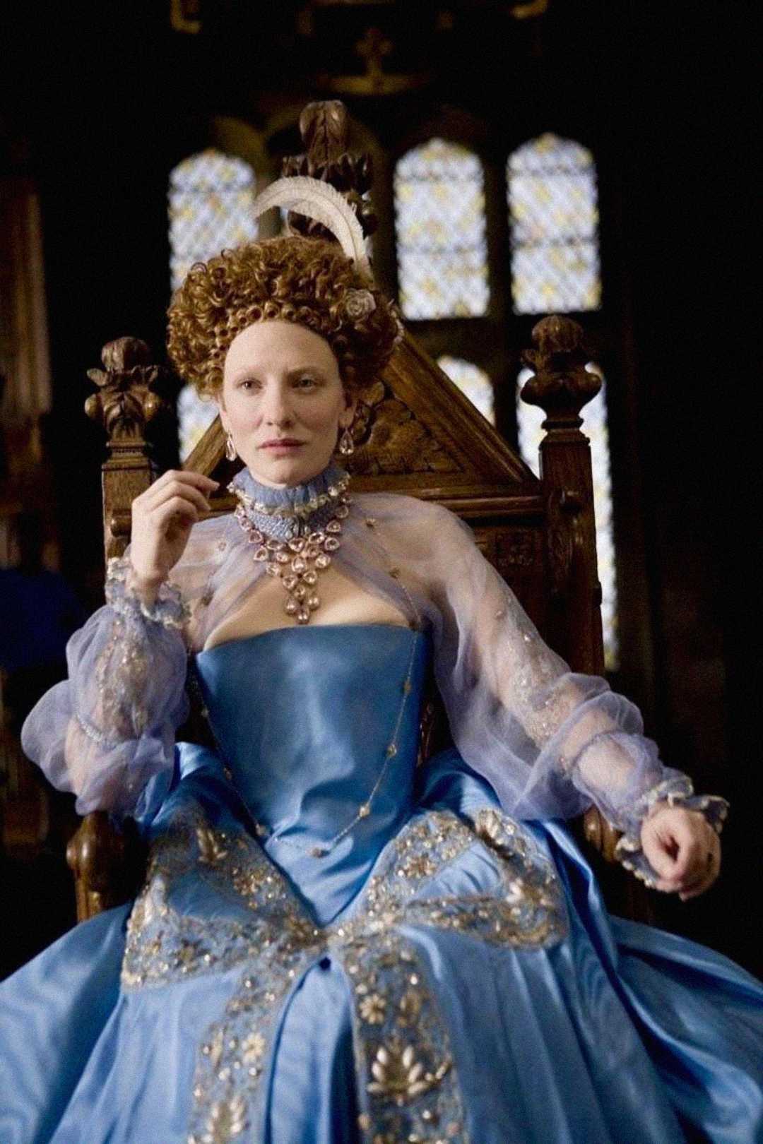 A woman in an ornate blue gown and jeweled necklace sits on a wooden throne, wearing an elaborate headpiece and sheer sleeves, against a background of stained glass windows.