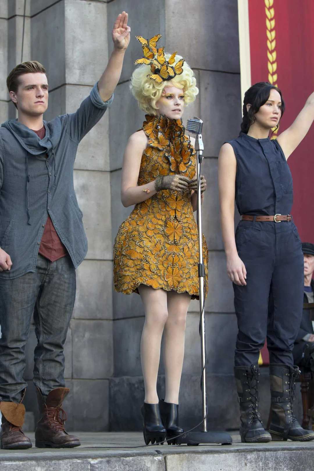 Three people stand on a stage; one woman in a yellow floral dress with a matching headpiece stands at a microphone, while a man and another woman beside her both raise their right arms in a salute.