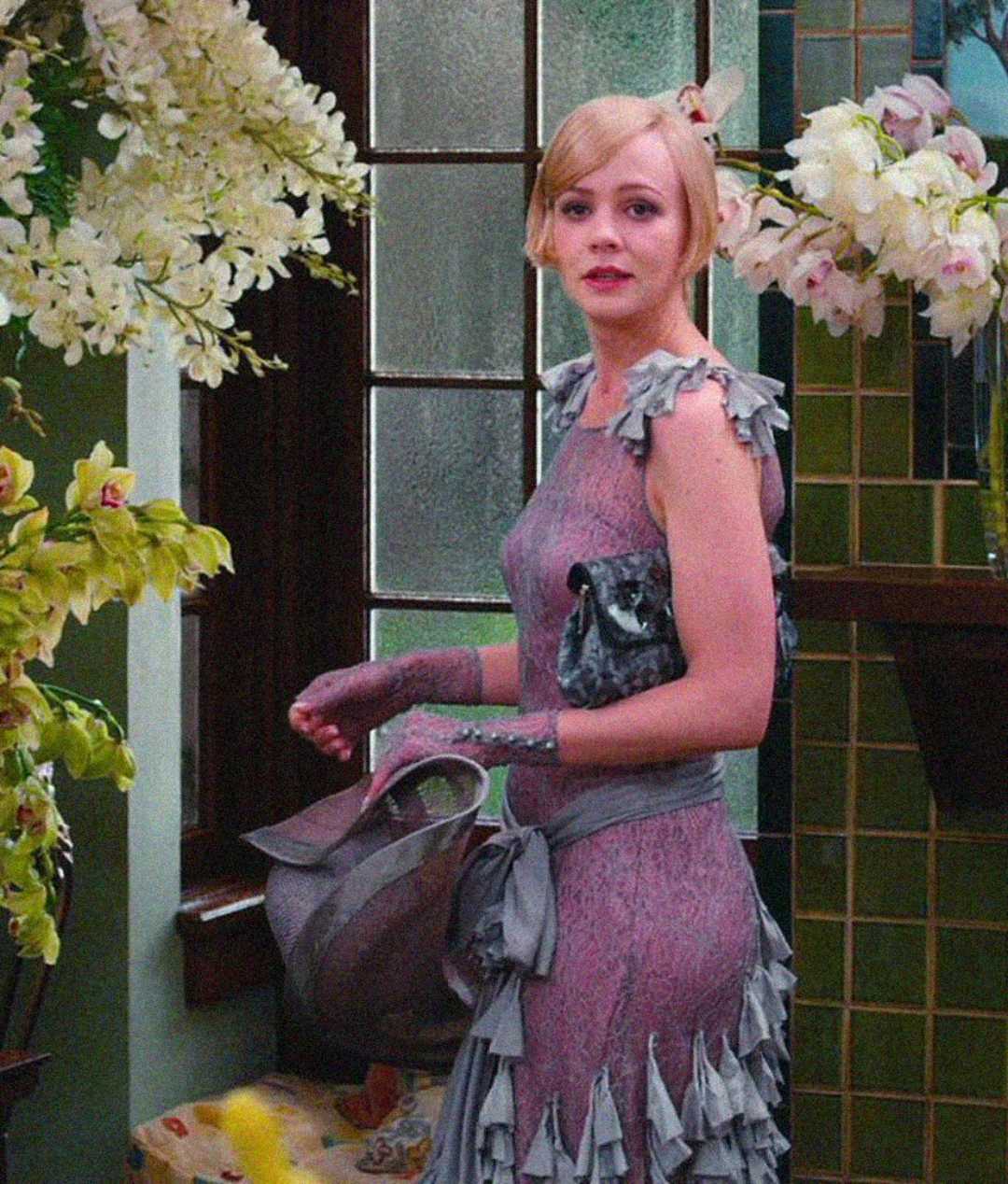 A woman in a lavender, vintage-style dress and gloves stands indoors, holding a large hat and a purse. She is surrounded by lush white flowers and green-tiled walls, looking slightly over her shoulder.