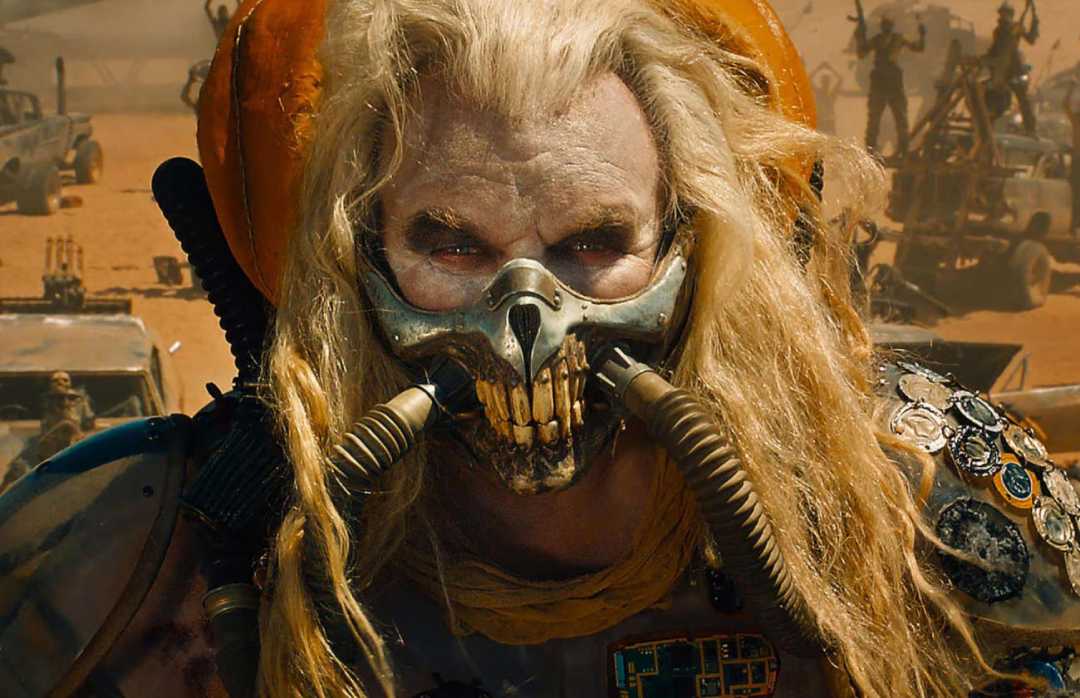 A person with long blond hair wears a menacing metal mask with tubes and armor in a post-apocalyptic desert setting, with vehicles and figures blurred in the background.