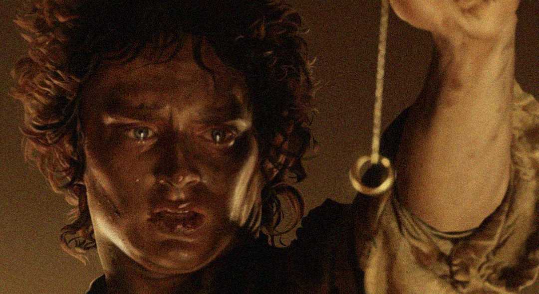 A person with curly brown hair, lit dramatically, looks intently at a gold ring hanging from a chain held up in their hand. The expression on their face is intense and focused.