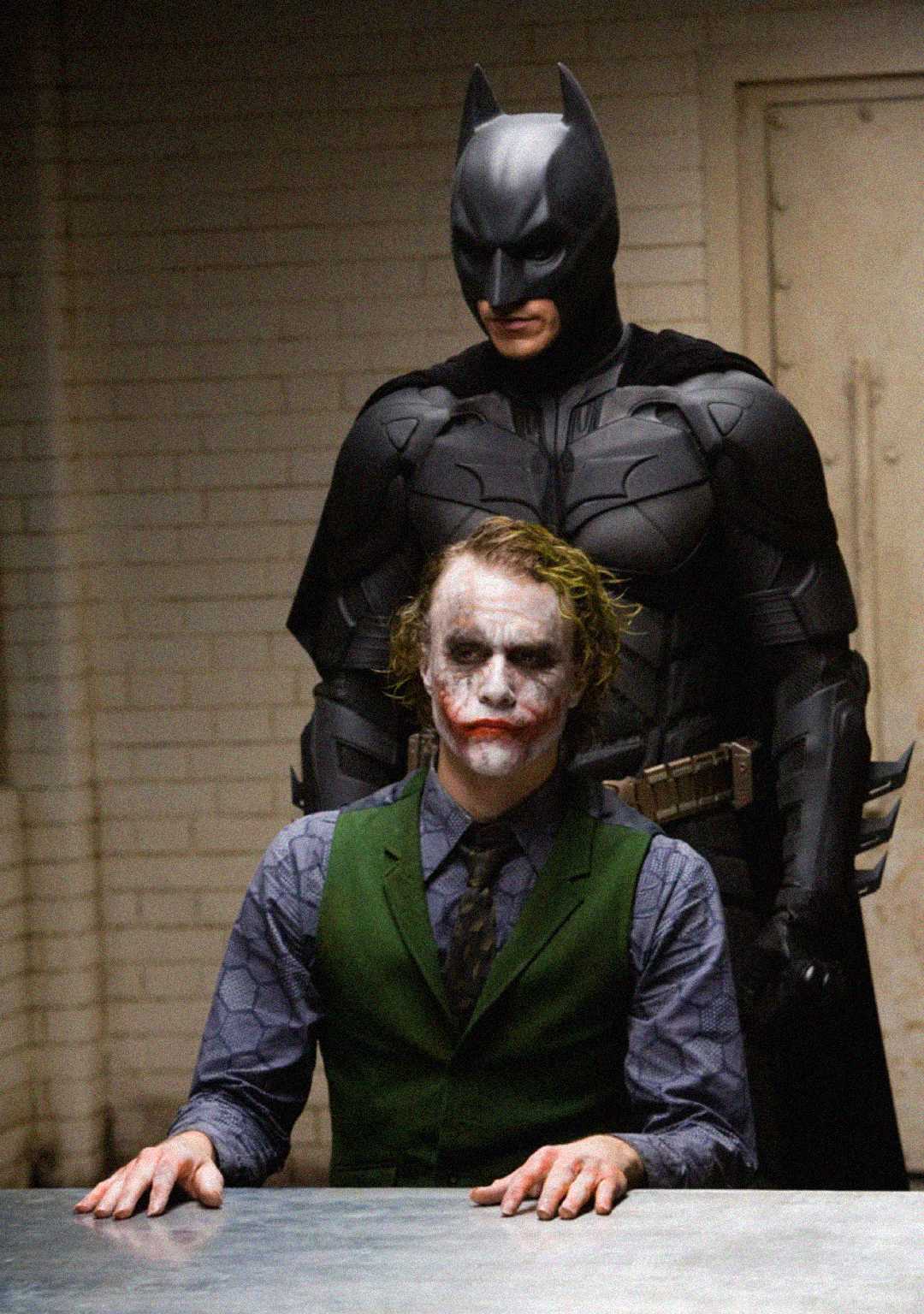 Batman stands behind the Joker, who is seated at an interrogation table in a dimly lit room. The Joker wears messy makeup and a green vest, while Batman is in his black armored suit.