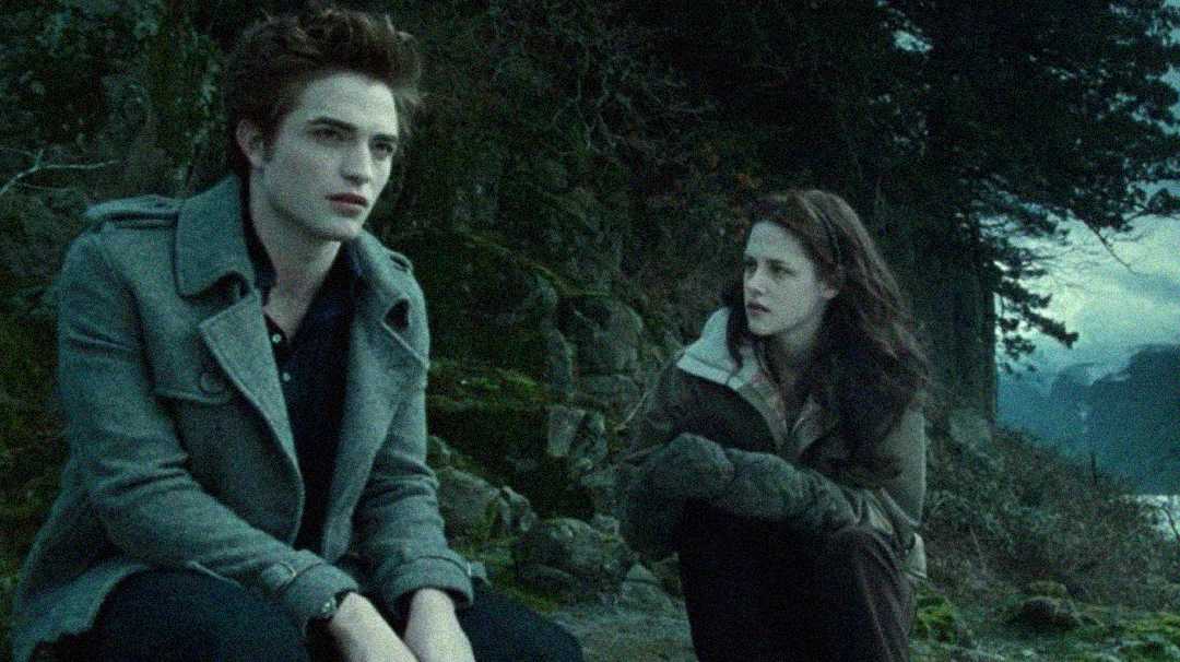 A young man and woman sit on rocks in a forested area near water, both looking serious. The scene has a cool, muted color tone, suggesting a somber or intense conversation.
