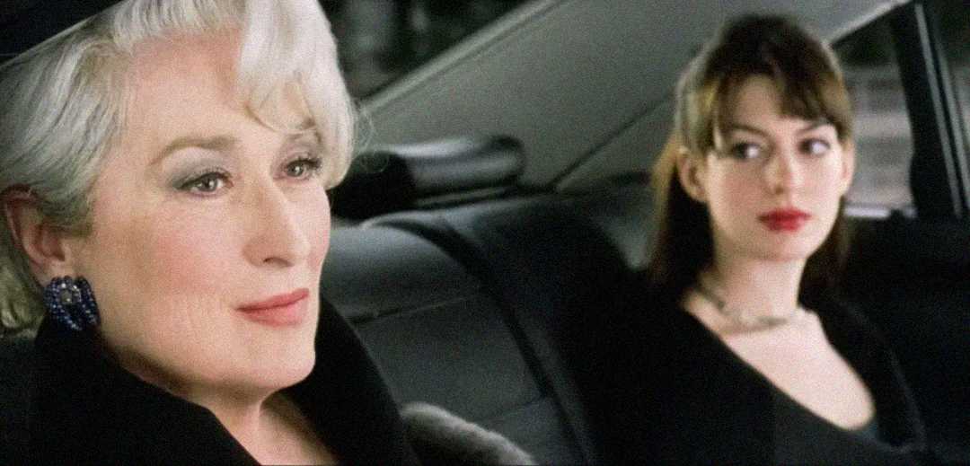Two women sit in the backseat of a car; one with short white hair looks ahead calmly, while the other with brown hair looks at her with a serious expression. Both are dressed formally.