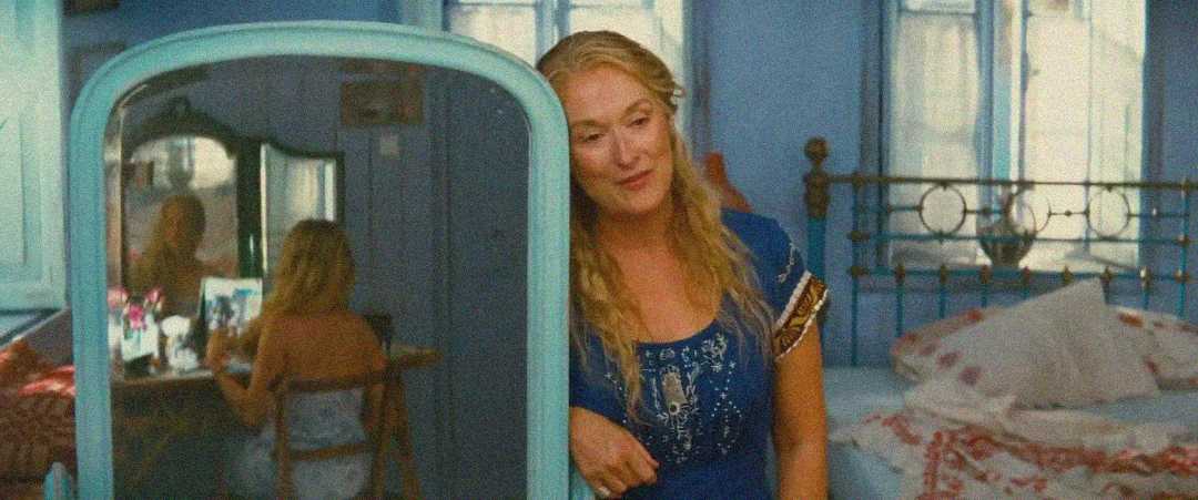 A woman in a blue dress leans against a blue wardrobe in a cozy, sunlit bedroom. Her reflection is visible in the mirror, while another woman sits at a vanity in the background. The room has blue walls and a brass bed.