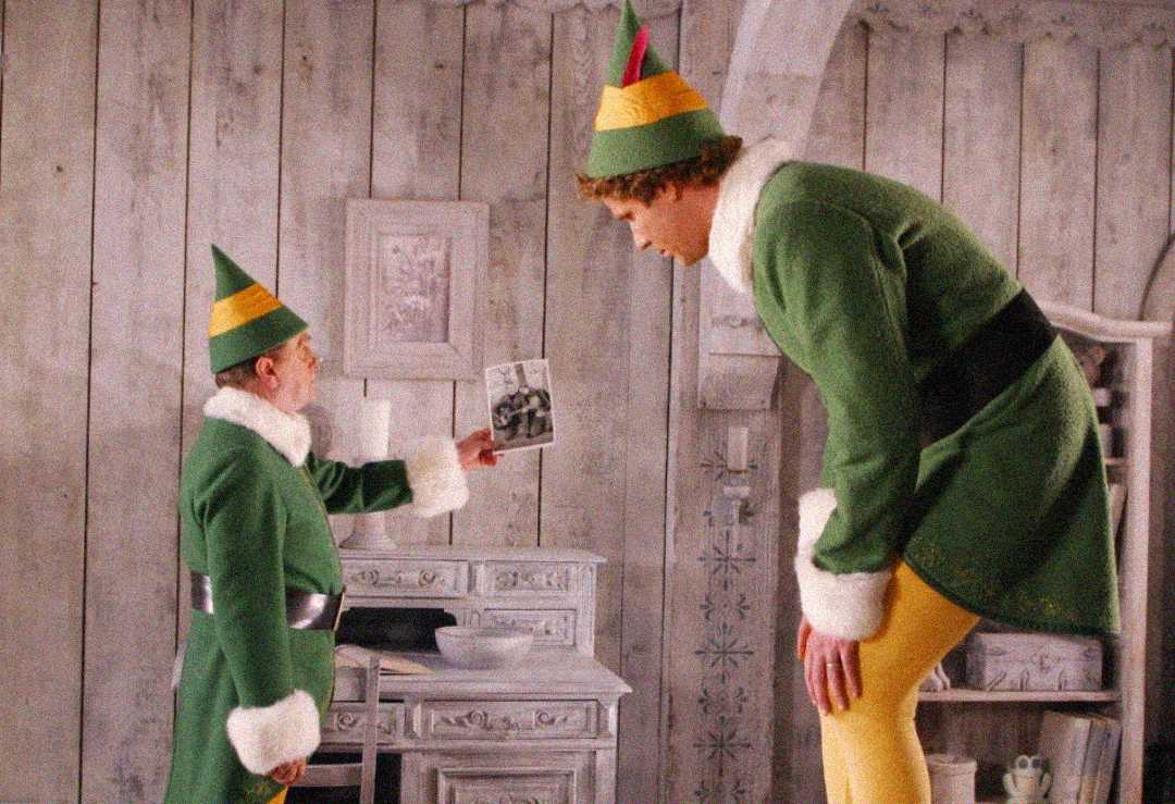 Two people dressed as elves stand in a rustic room. One, much shorter, holds up a photograph to the taller elf, who is bending over to look at it. Both wear green costumes with yellow hats and white trim.