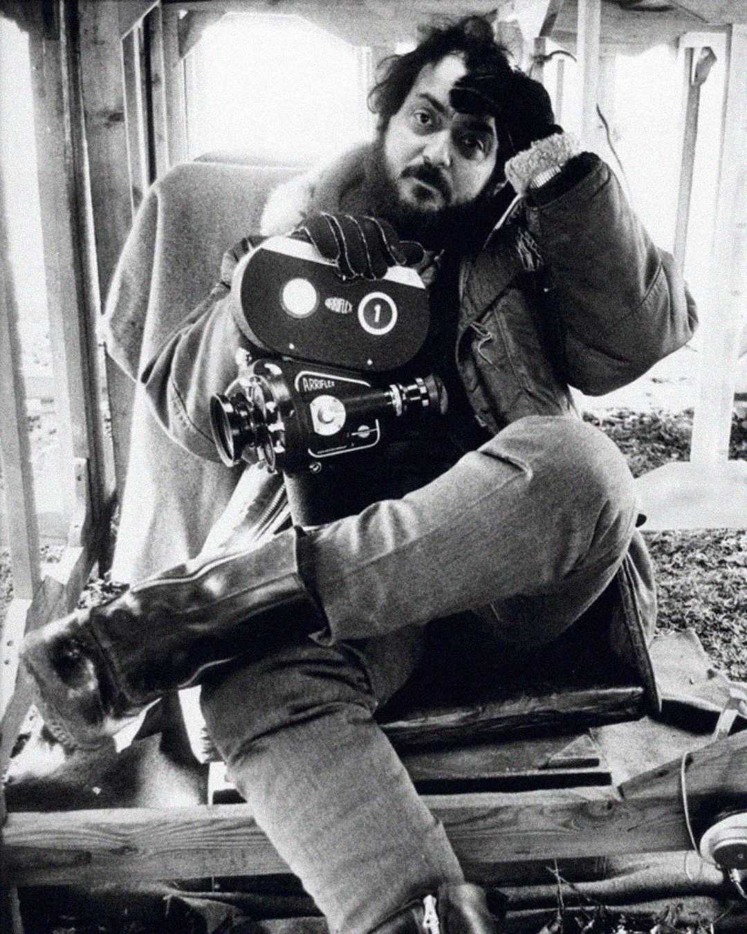 A bearded man in a heavy jacket and boots sits cross-legged, holding his head with one hand and a vintage film camera with the other, inside a wooden structure. The image is in black and white.