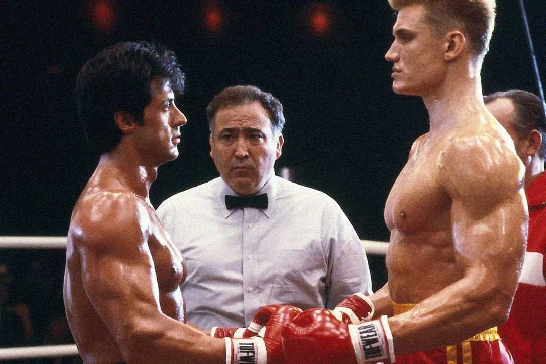 Two muscular boxers face each other in a boxing ring, both wearing gloves and shorts, with a referee standing between them, looking serious. The background is dimly lit with visible spectators.