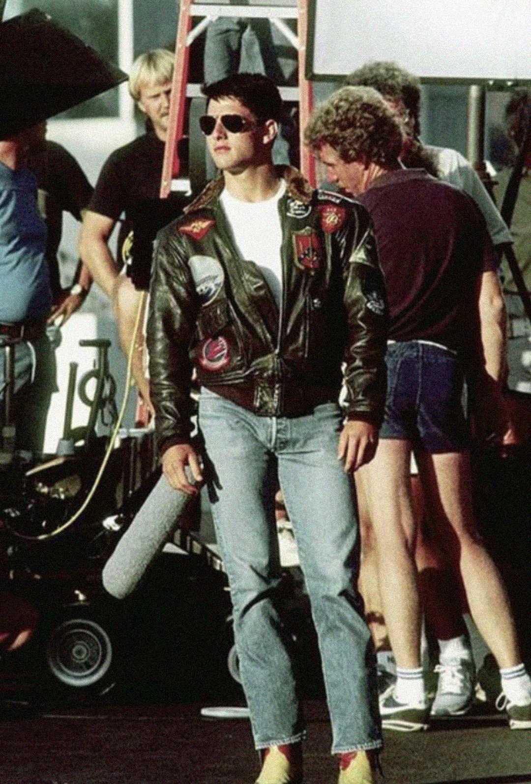 A man in a leather flight jacket, white T-shirt, jeans, and sunglasses stands confidently on a film set, surrounded by crew members and equipment in the background.