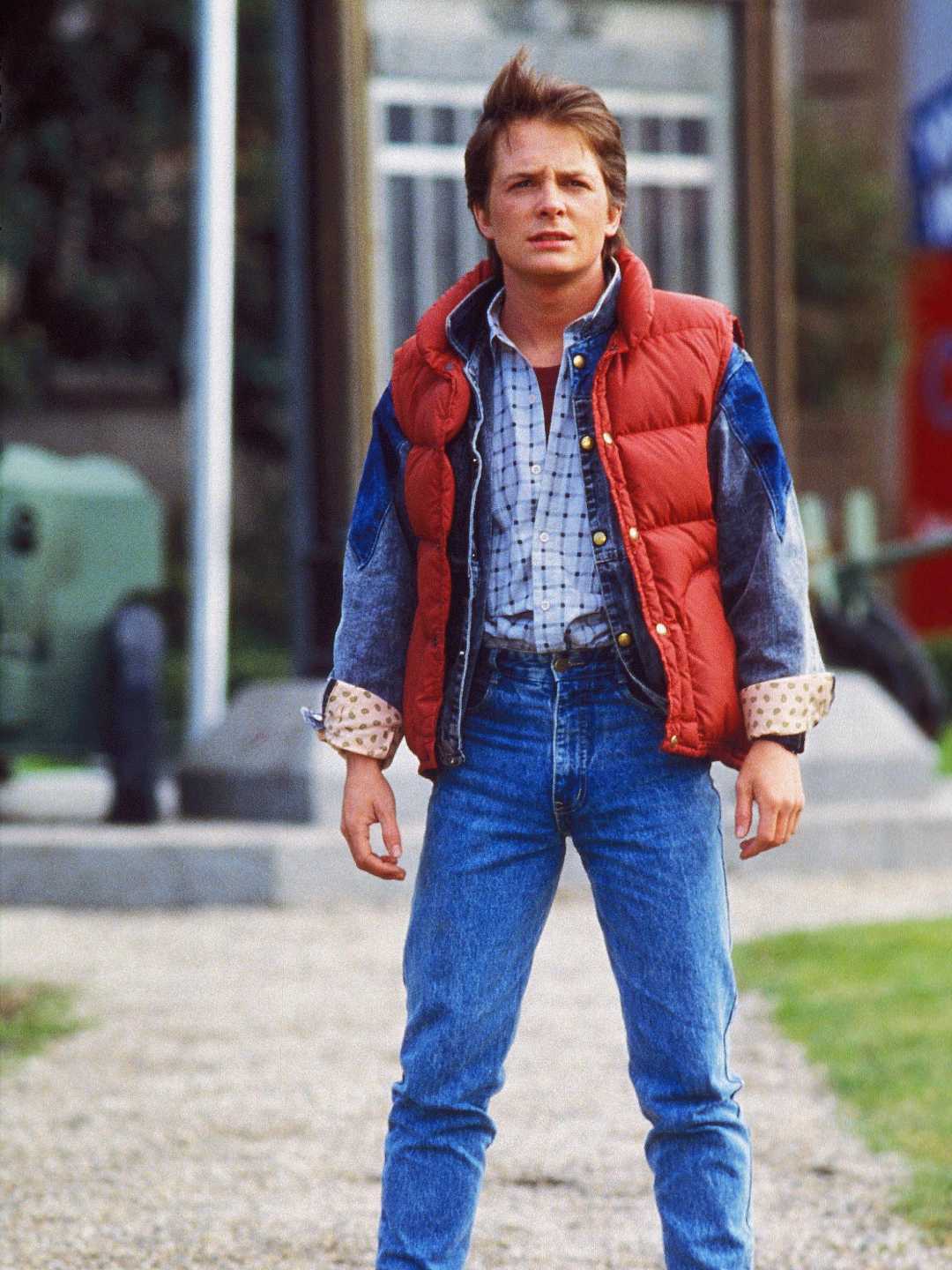A young man stands outdoors wearing a red puffy vest over a denim jacket, plaid shirt, and blue jeans. He has short brown hair and looks off into the distance with a serious expression.