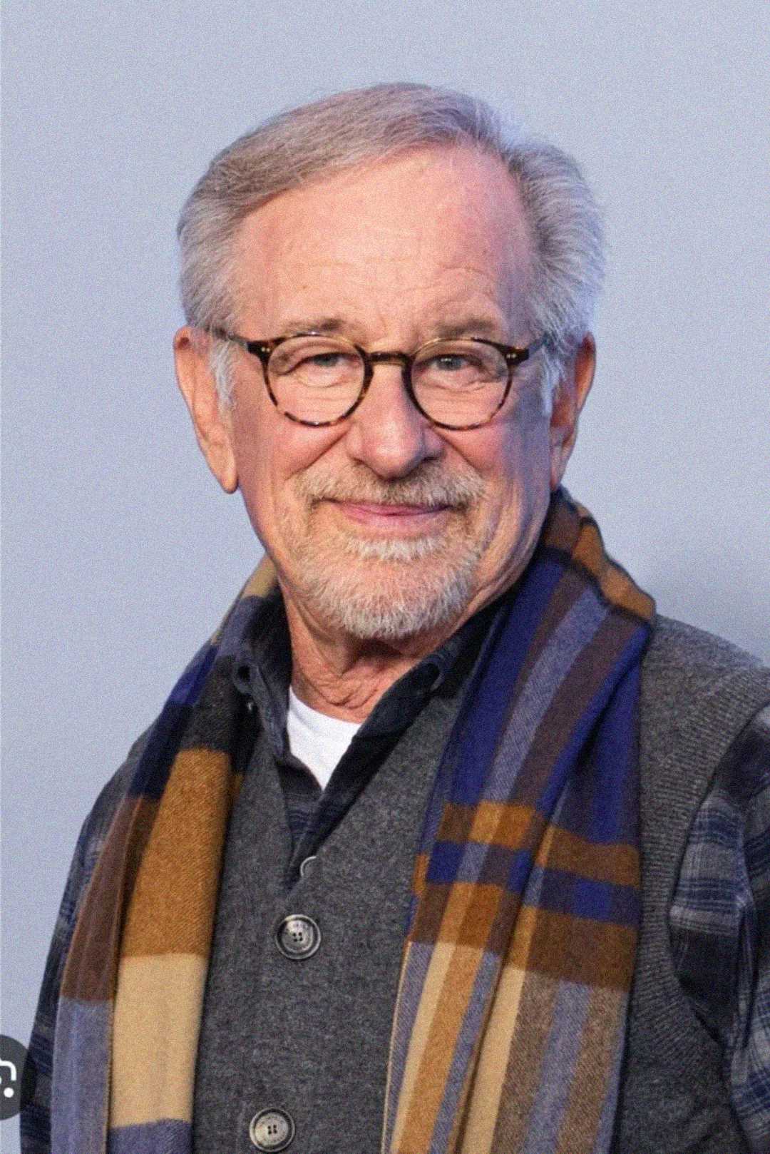 An older man with gray hair, glasses, and a short beard smiles while wearing a gray sweater, plaid shirt, and a blue and brown scarf. The background is plain and light-colored.