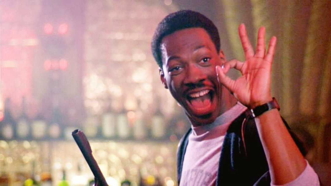 A man smiling widely makes an "OK" gesture with his hand, holding a broom and standing in a bar with bottles visible in the blurred background.