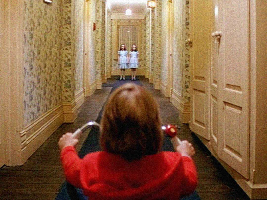 A child on a tricycle rides down a dimly lit hallway toward two identical girls standing side by side at the end, in a scene with floral wallpaper and closed doors lining the corridor.