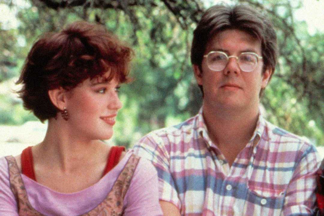 A young woman with short red hair looks at a young man with glasses and dark hair; they are sitting outdoors, and the man is wearing a plaid shirt while the woman smiles at him.