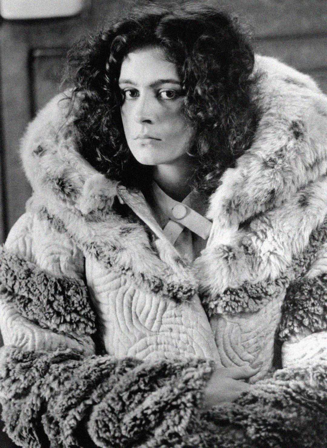 A young woman with curly hair wears a large, textured fur coat. She looks slightly to the side with a serious expression. The black-and-white photo has a vintage feel.