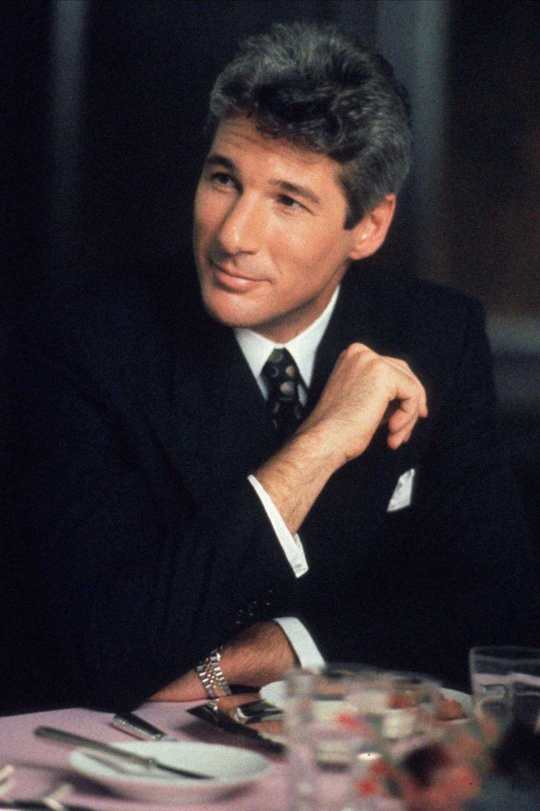 A man in a dark suit and tie sits at a dining table, resting his chin on his hand and looking slightly to the side with a gentle smile. He has short, gray hair and there are plates and glasses on the table.