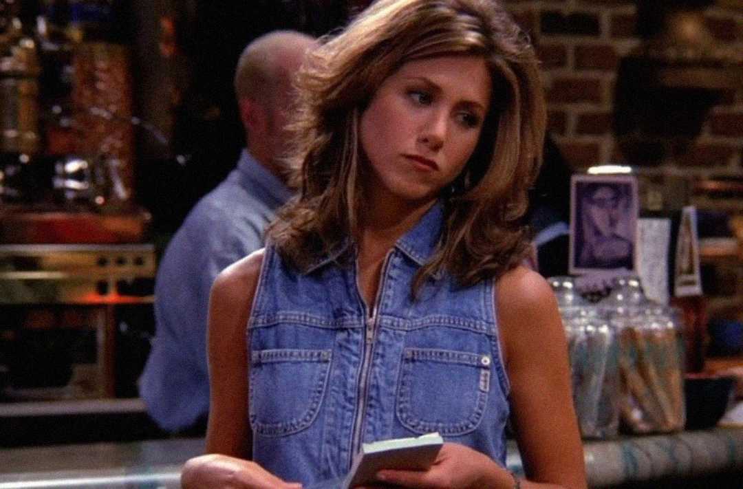 A woman with layered brown hair, wearing a sleeveless denim shirt, stands indoors holding a notepad, looking to the side with a neutral expression. The background is blurred with brick walls and cafe items visible.