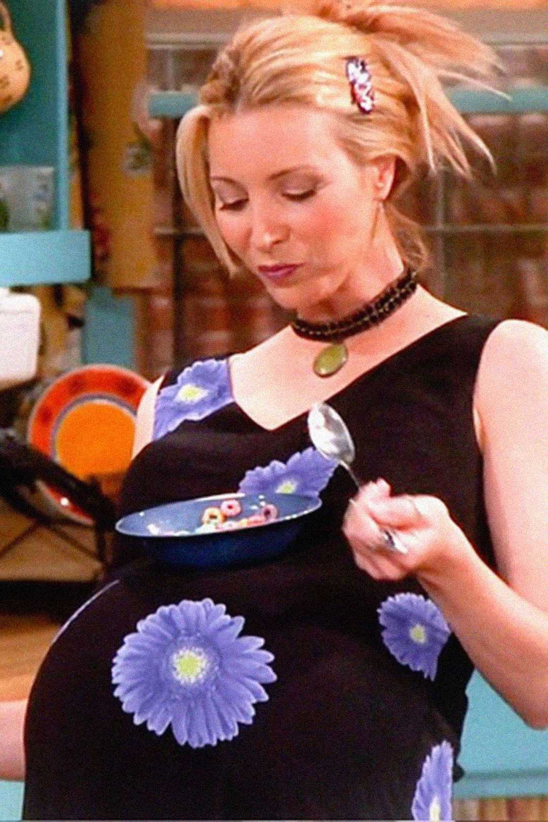 A pregnant woman with blonde hair in a black dress with blue flowers balances a blue bowl on her belly and eats cereal with a spoon, smiling slightly. The background shows a colorful kitchen.