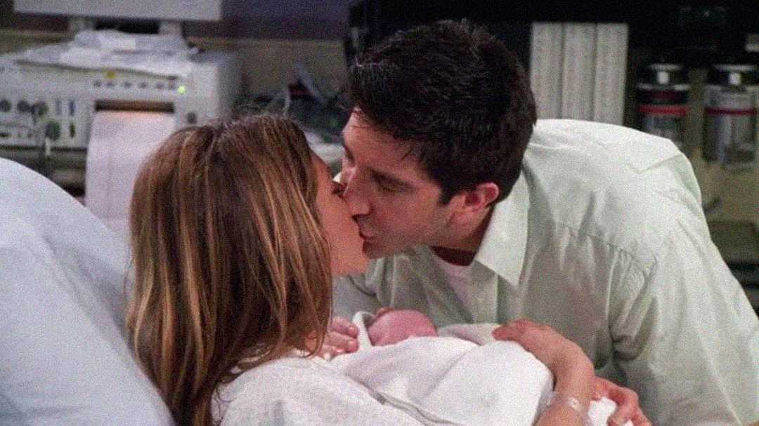 A woman lying in a hospital bed holds a newborn baby while kissing a man who leans over her. Hospital equipment is visible in the background, suggesting a maternity ward setting.