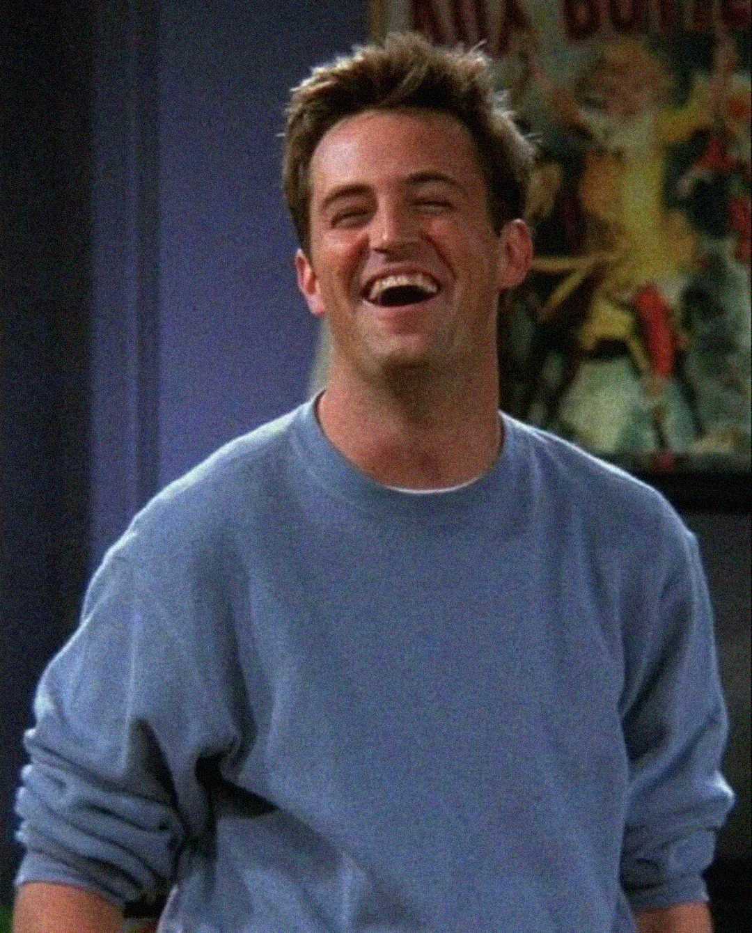 A man with short brown hair wearing a light blue sweatshirt is laughing, standing indoors with a blurred colorful poster in the background.