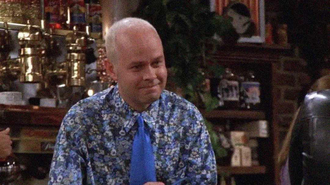 A man with a bald head wears a blue floral shirt and matching tie, standing behind a counter in a coffee shop with shelves and equipment in the background.