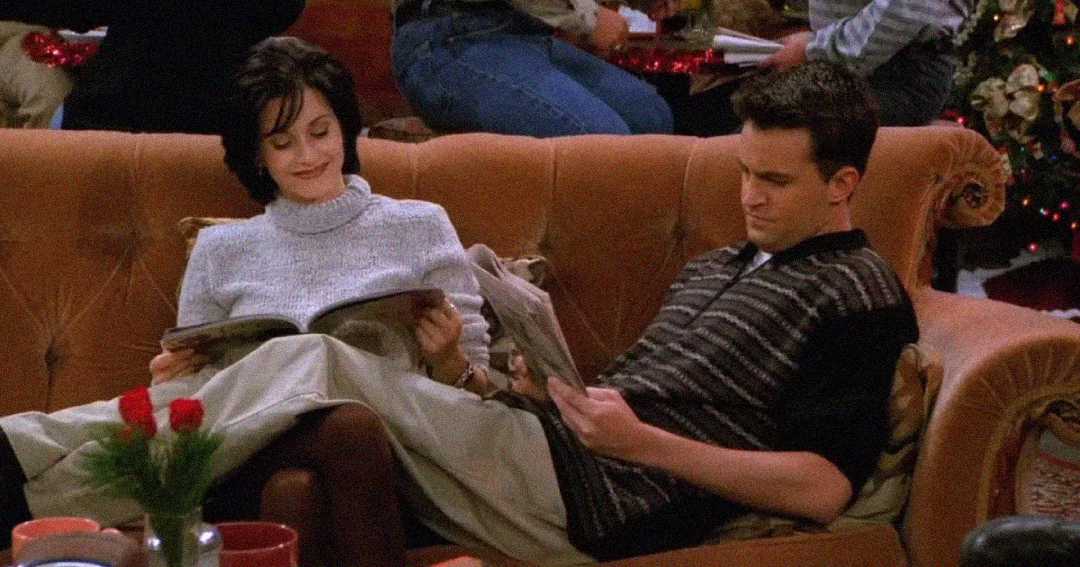 A woman and a man sit closely together on an orange couch, each reading a magazine. The setting appears cozy, with people and a decorated Christmas tree in the background.