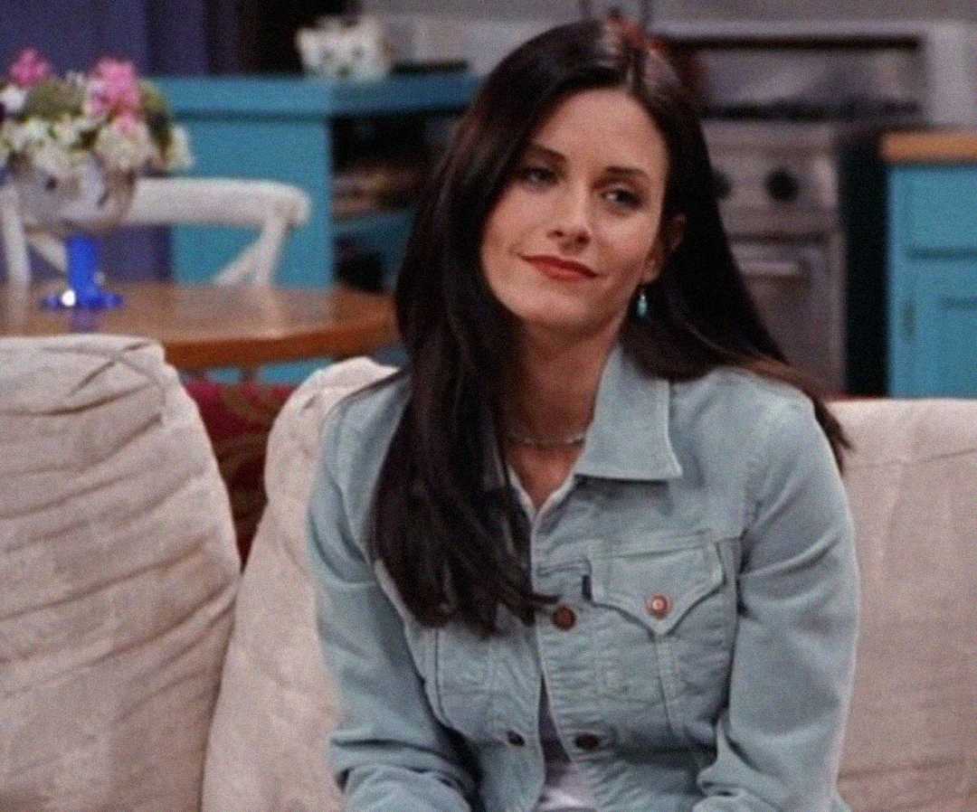A woman with long dark hair, wearing a light blue denim jacket, sits on a beige couch in a cozy living room with blue cabinets and a vase of flowers in the background. She has a slight, amused smile on her face.