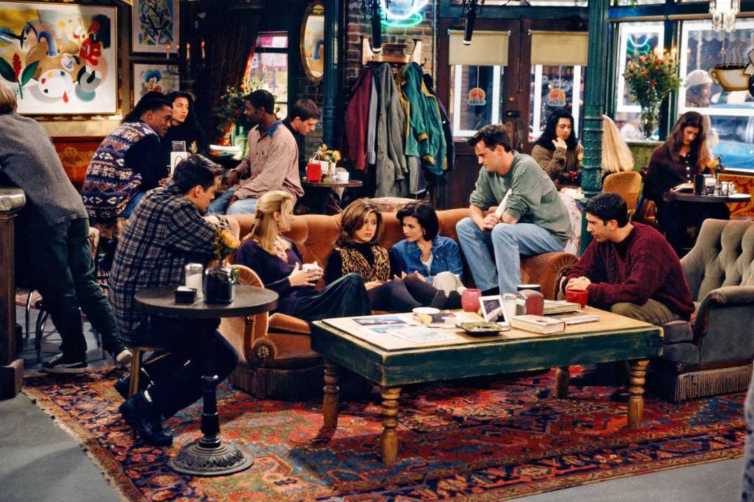 Six friends sit and talk on sofas and chairs around a coffee table in a cozy, busy coffee shop with people in the background, colorful decor, and a large window letting in daylight.