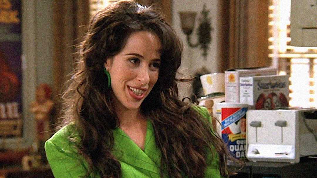 A woman with long, wavy brown hair, wearing a bright green jacket, smiles and tilts her head in a room with shelves, boxes, and various items in the background.