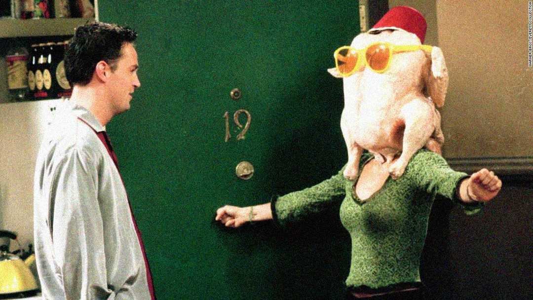 A person wearing a green sweater stands with arms outstretched and a raw turkey on their head, decorated with sunglasses and a red hat, while another person in a light shirt looks on near a green door labeled "19.