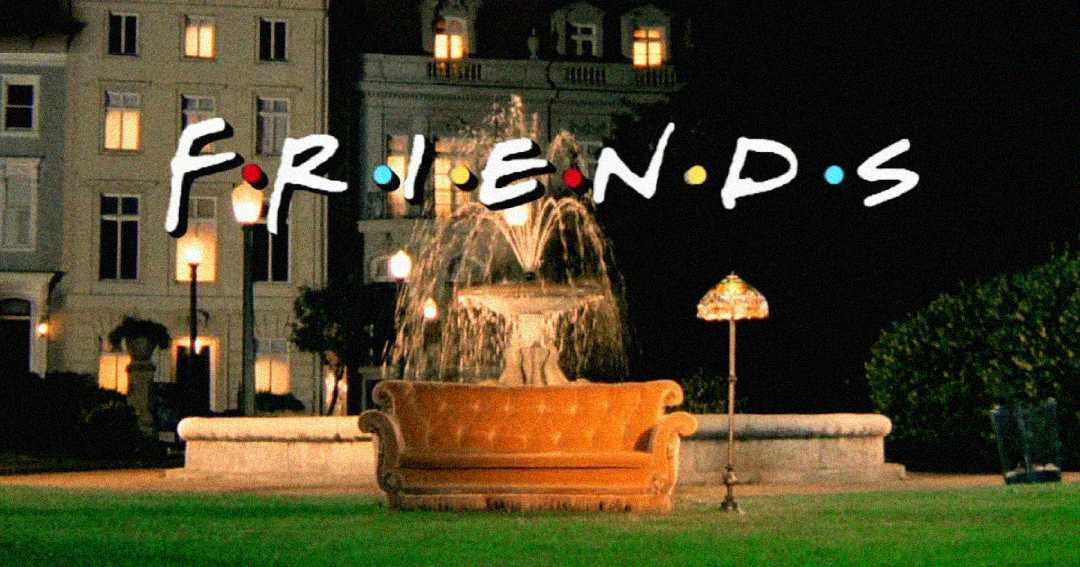 A brown couch sits in front of a lit fountain at night, with the word "FRIENDS" above it in white letters, each letter separated by colorful dots. Buildings and lampposts are visible in the background.