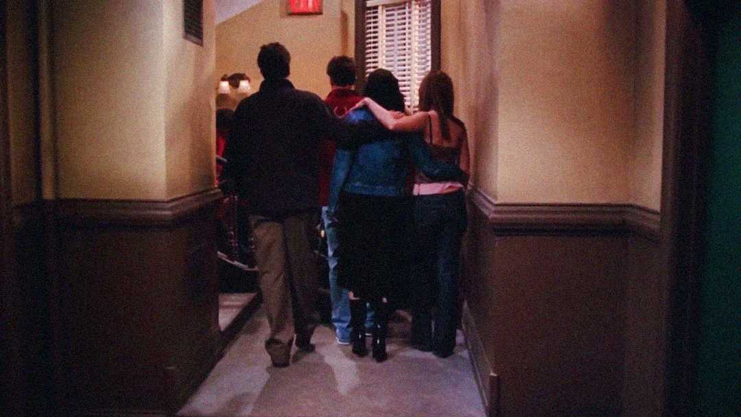 Four people are walking down a dimly lit hallway with their arms around each other’s shoulders, viewed from behind. They appear to be leaving together, creating a sense of friendship and togetherness.